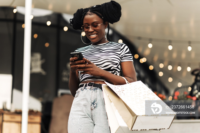 Young african woman, in a mall using a mobile phone, with bags makes purchases for Christmas