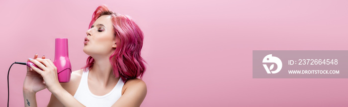 young woman with colorful hair blowing at hairdryer isolated on pink, panoramic shot