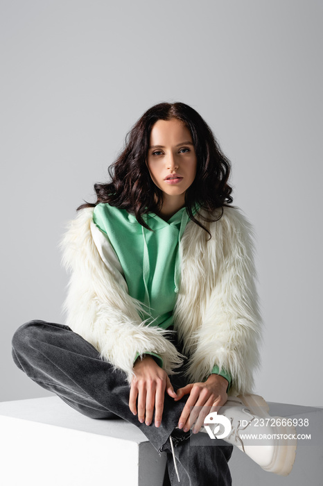 young woman in faux fur jacket posing on cube on white background