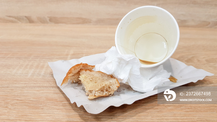 Leftover food on a paper plate and empty dirty paper coffee cup on a wooden table in a fast food caf