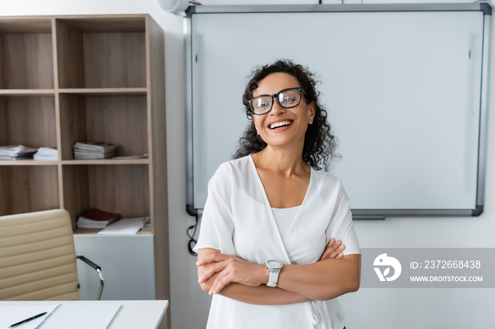 happy african american teacher in eyeglasses standing with crossed arms in classroom