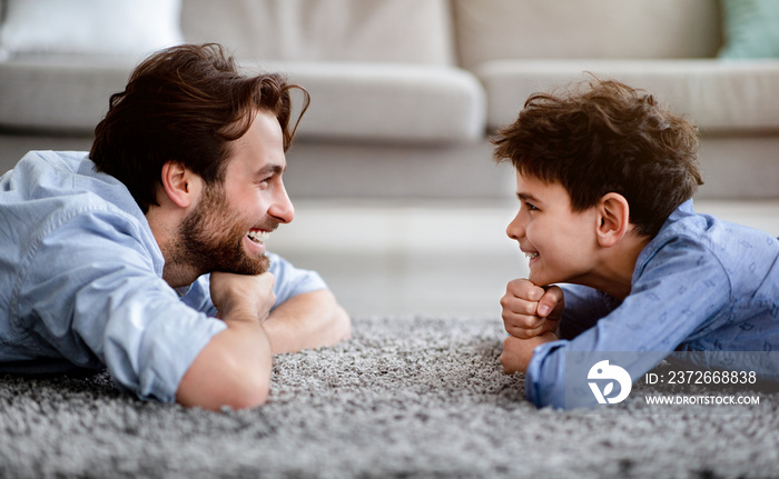 Two generations. Profile portrait of happy father and his son lying on carpet at home and looking at