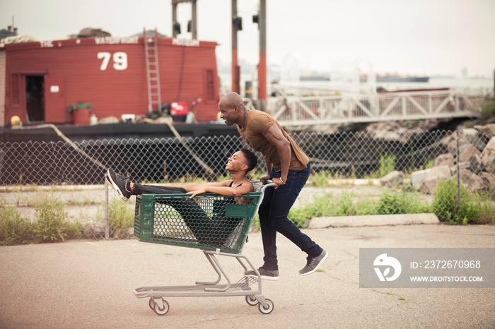 Side view of boyfriend pushing girlfriend sitting in shopping cart on road at parking lot