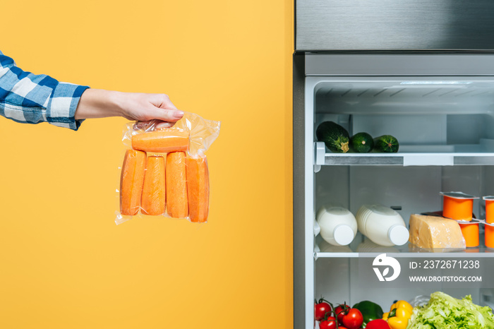 打开的冰箱附近，一名妇女拿着真空包装中的胡萝卜，货架上有新鲜食物的裁剪图