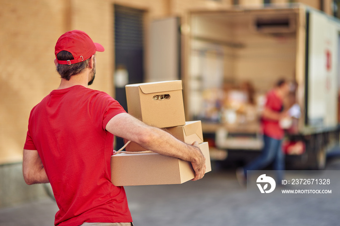 Rear view of a male courier in red uniform holding parcels for delivery while standing at storehouse