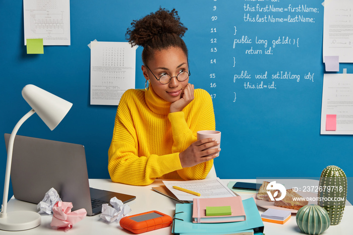 Contemplative Afro female freelancer drinks caffeine beverage from cup, looks pensively aside, dress