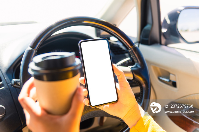 一名亚洲女子在车内喝热咖啡外卖杯，在开车时使用智能手机空白屏幕