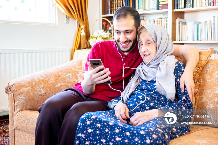 Happy arabic muslim grand mother and her son sitting together on sofa and using mobiles