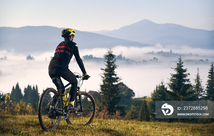 穿着骑行服的男子在长满草的小山上骑自行车的后视图。戴着安全帽的男自行车手enjo