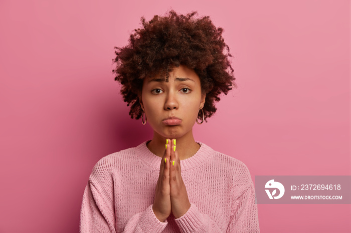 Hopeful sad uneasy curly woman beggs for forgiveness, purses lips and keeps palms in praying gesture