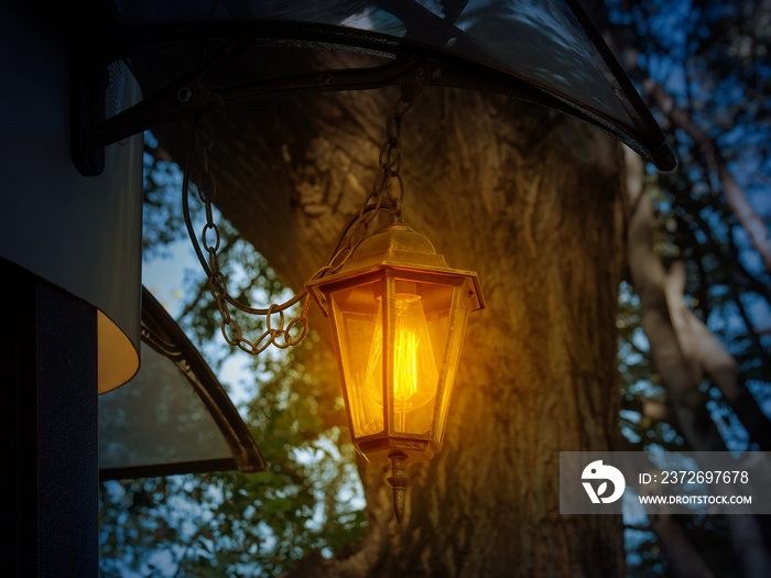 Retro style lantern shines with warm yellow light at dusk. Vintage lantern hangs on a chain under th