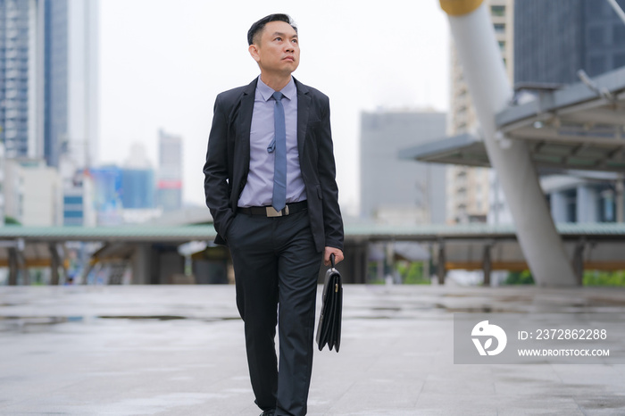 Asian Businessman walking and holding briefcase with business office buildings in the city backgroun