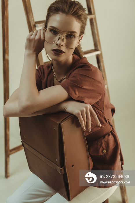 beautiful stylish girl holding leather bag and sitting near ladder isolated on beige