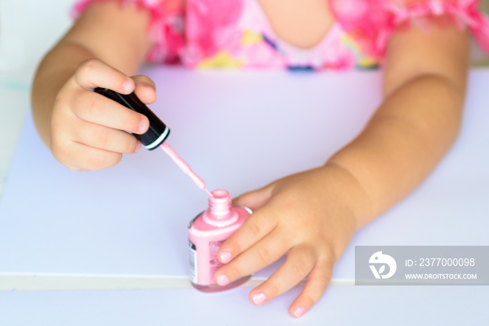 Adorable little girl having fun playing at home with colorful pink nail polish doing manicure and painting nails. Toddler cute child in pink elegant dress sitting wood white table and making manicure