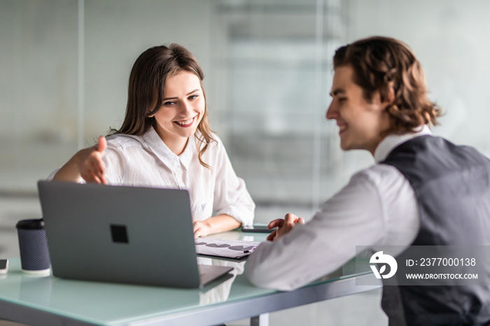 Beautiful young business woman and handsome businessman in formal suits are using a laptop, discussing documents and smiling while working in office