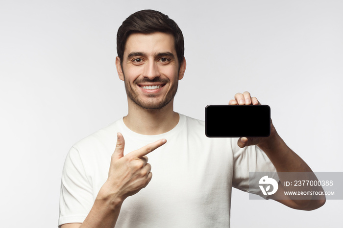 Happy young man pointing with finger to blank smartphone, smiling at camera, isolated on gray background