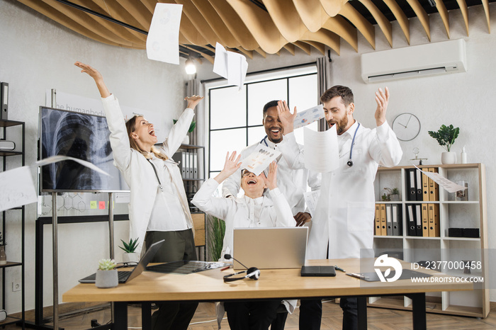 Excited multiracial scientists in white medical uniform laughing and throwing documents in air during laboratory meeting. Success teamwork and healthcare achievement concept.