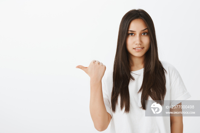 What do you know about that person. Portrait of questioned attractive female coworker in white t-shirt, pointing left with thumb, lifting eyebrow curiously and smiling broadly, asking question