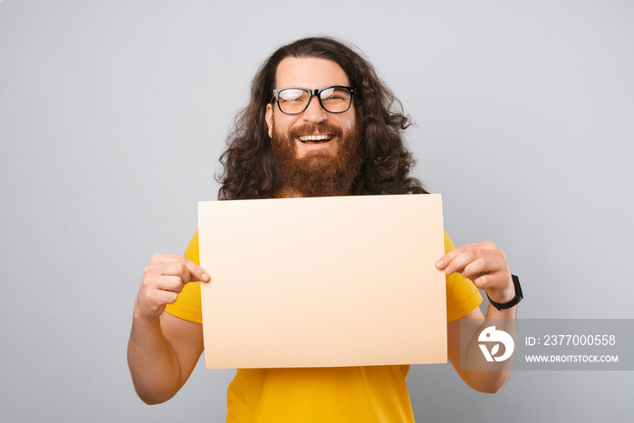 Wide smiling handsome man is holding a piece of paper with a message for you. Portrait over gray background.