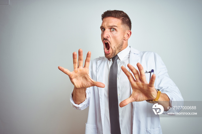 Young handsome doctor man wearing white profressional coat over isolated background afraid and terrified with fear expression stop gesture with hands, shouting in shock. Panic concept.