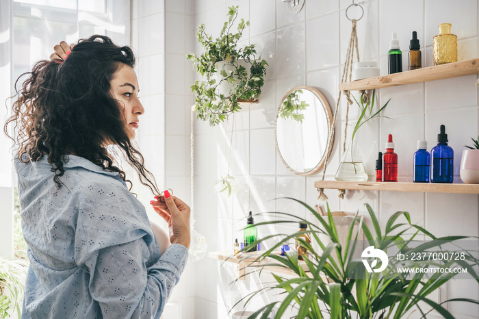 Young overweight brunette woman with curly hair looking into mirrow in light bathhroom with wooden shalves for natural cosmetics. Wellness and body positivity concept