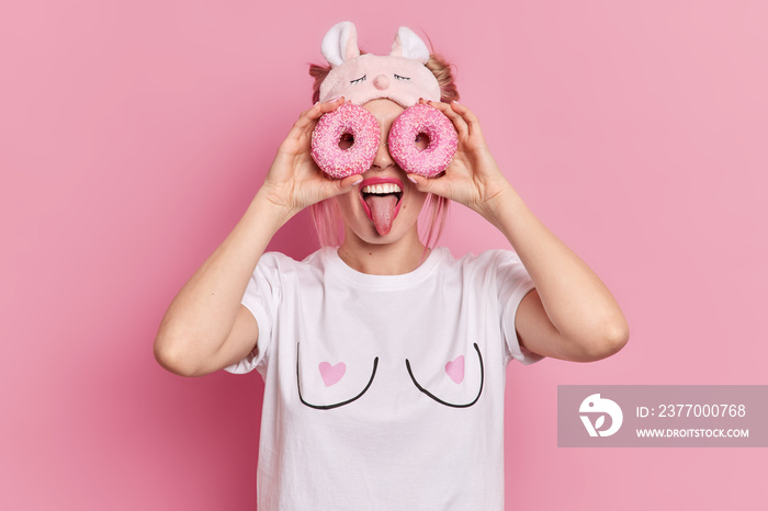 Funny woman covers eyes with two glazed donuts starves for sweets sticks out tongue has sweet tooth has playful mood wears blindfold and white t shirt isolatd over pink background. Junk food