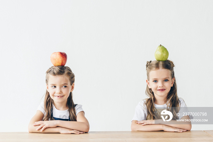 adorable little schoolgirls with apple and pear on heads sitting at table isolated on white