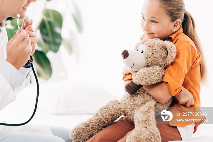 partial view of pediatrist in white coat and kid with teddy bear in clinic