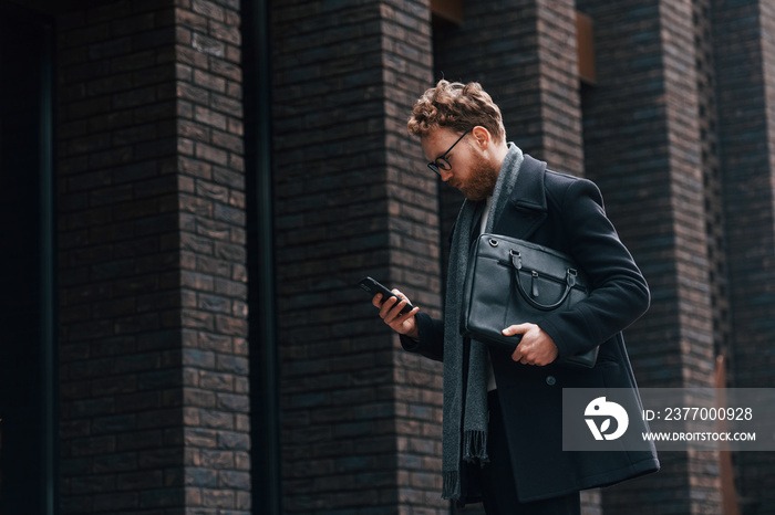 Holding smartphone. Stylish man with beard and in glasses is outdoors near building