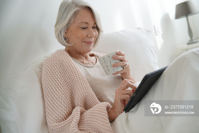 Attractive senior woman relaxing in bed with hot drink and tablet