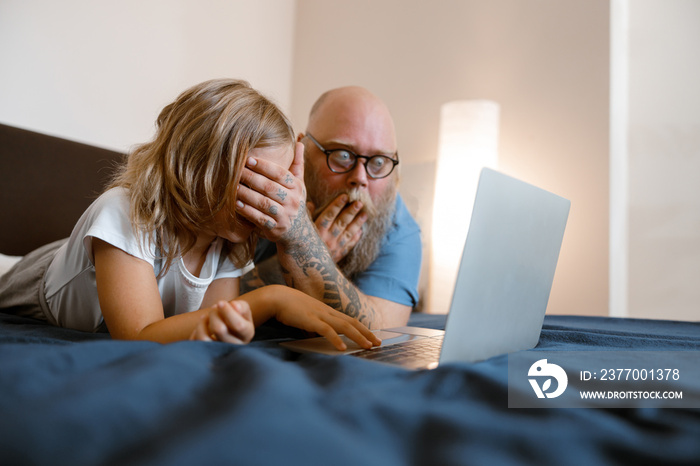 Shocked father closes eyes of daughter to protect from dangerous content on laptop on bed