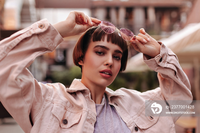 Short haired woman in light jacket and pink glasses looking into camera outdoors. Trendy brunette lady in beige clothes posing in city..