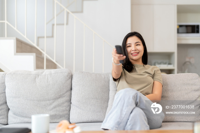 Image of a beautiful asian woman searching channel with remote control to watch tv while sitting on sofa at home