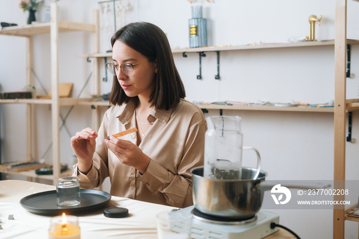 Medium shot of attarctive female artisan using piece of beeswax to create wick for handmade candle at workshop. Process of making hand-made natural candle at home.