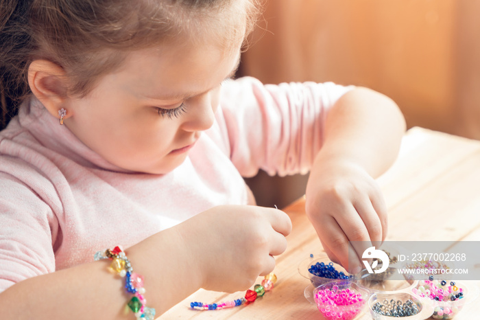 A little girl is engaged in needlework, making jewelry with her own hands, stringing multi-colored beads on a thread