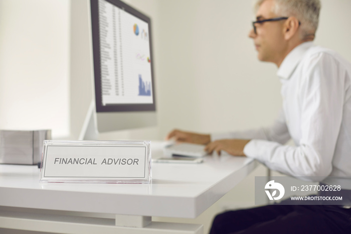 Closeup nameplate sign plaque board that reads Financial Advisor and male professional using computer in background. Senior worker sitting at office desk and looking at business data graphs and charts