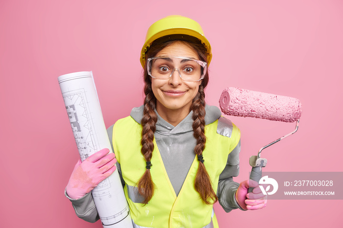 Positive woman maintennce worker checks construction draft uses paint roller for remodeling house helps clients to redecorate room wears protective hardhat transparent glasses reflecting vest