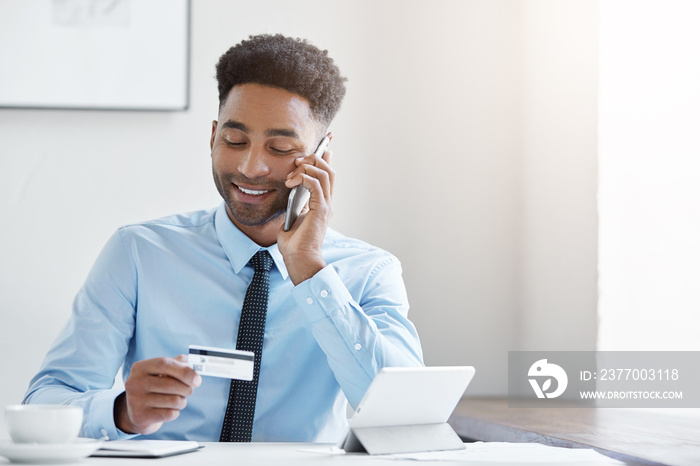 Young mixed race businessman wearing official clothes, sitting at spacious office, paying bill with credit card, using his modern smart phone. Professional young business worker with credit card