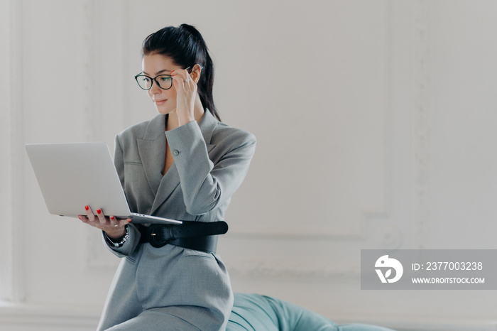 Elegant professional woman banker concentrated at screen of laptop computer, dressed in formal wear, reads documents in online version poses in spacious room, home office. Business, technology concept