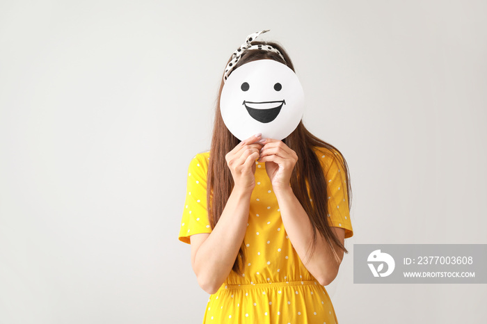 Woman hiding face behind sheet of paper with drawn emoticon on grey background