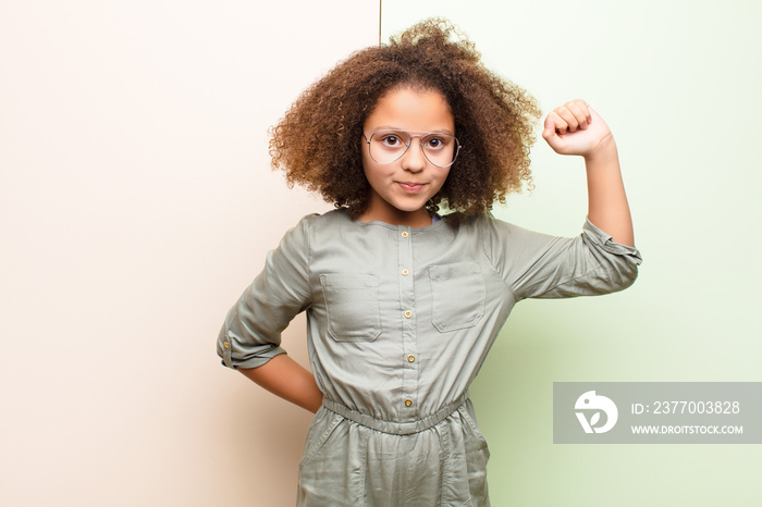 african american little girl feeling serious, strong and rebellious, raising fist up, protesting or fighting for revolution against flat wall