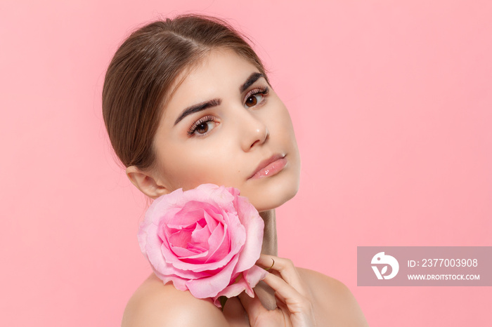 Close-up portrait of a beautiful young girl looking at the camera holding pink rose flower close to face isolated over pink background.