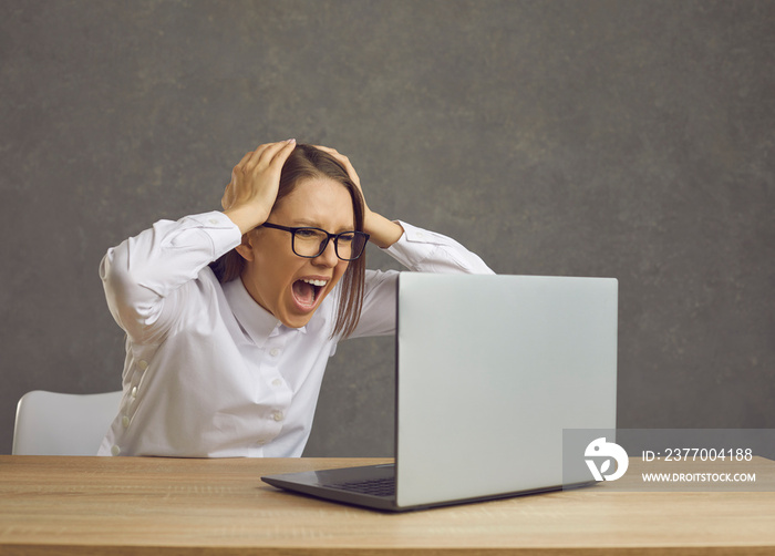 Portrait of young angry shocked woman in eyeglasses looking at laptop computer screaming grabbing head. Female businesswoman freelancer emotionally showing facial expressions sitting at desk