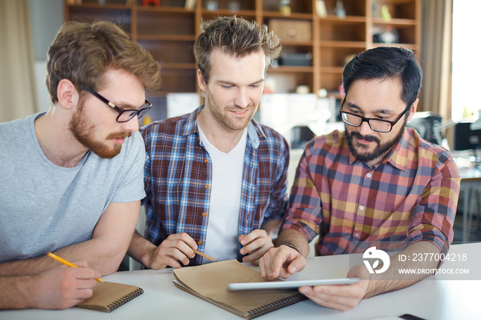 Team of young entrepreneurs at work:  three young ambitious men sitting at table planning strategy to new business idea, Asian man pointing at tablet screen, the other two ready to make notes