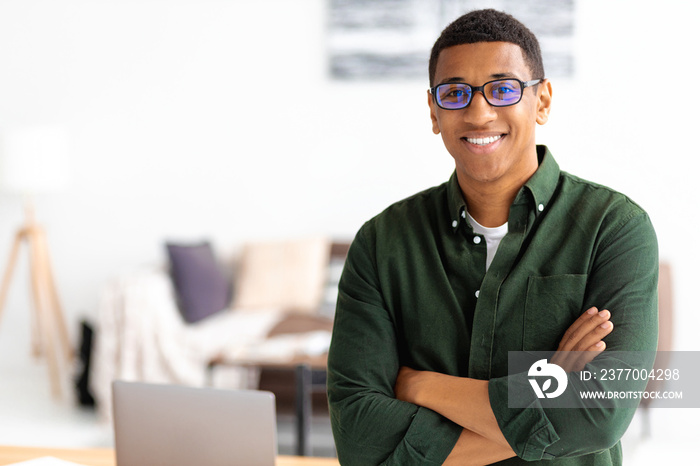 Smiling successful businessman with eyeglasses stands with crossed arms smiling and looking at the camera. Young male confident entrepreneur or small business owner in office, copy space