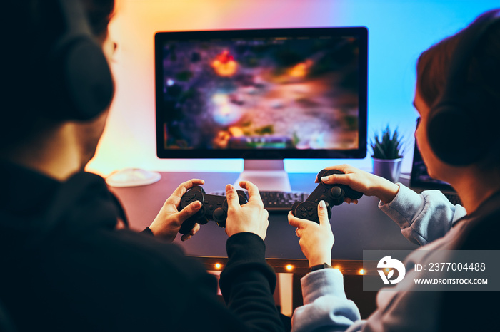 Friends playing video game at home. Gamers holding gamepads sitting at front of screen. Streamers girl and boy playing online in dark room lit by neon lights. Competition and having fun