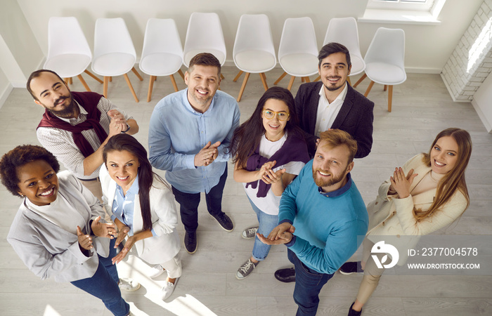 Diverse team of happy business people standing together and applauding. Group portrait of young male and female colleagues looking up at camera, smiling and clapping hands. High angle, shot from above