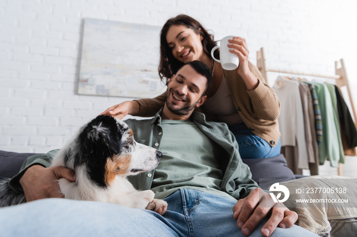 smiling young woman holding cup near bearded boyfriend hugging australian shepherd dog.