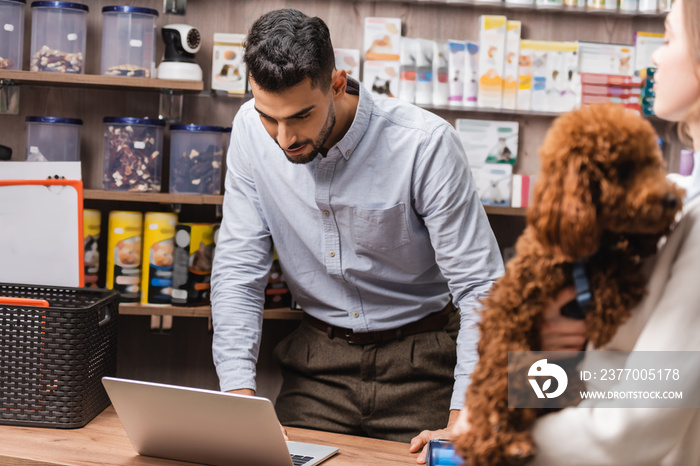 Arabian salesman looking at laptop near blurred customer with poodle in pet shop