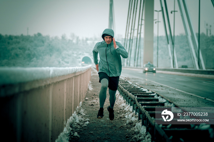 runner in motion fast running over the bridge at the winter snowy weather. Sportsman workout on cold weather. Athlete with hoodie in the sportswear.